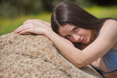 Portrait of a young brunette woman, with pale skin, wearing a blue top, shy and reserved, looking at the camera, sitting on a huge stone in an outdoor park.の写真素材