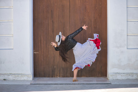 Young woman, beautiful, brunette, flamenco dancer, wearing a beautiful dress with hat, dancing flamenco with a wooden door in the background. Flamenco concept, typical, Spanish, Seville.の写真素材