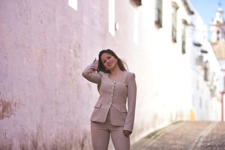 Young, pretty, brown-haired woman, wearing a light gray jacket suit, with one hand in her hair, walking down a lonely, Mediterranean-style street in a village.の写真素材