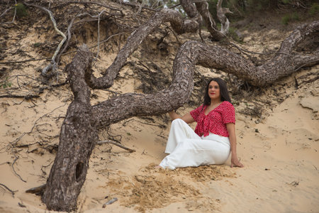Young, beautiful, brunette woman in red shirt and white pants, looking lost, sitting relaxed and happy on the sand next to a fallen log. Concept beauty, relax, nature, calm, happiness.の写真素材