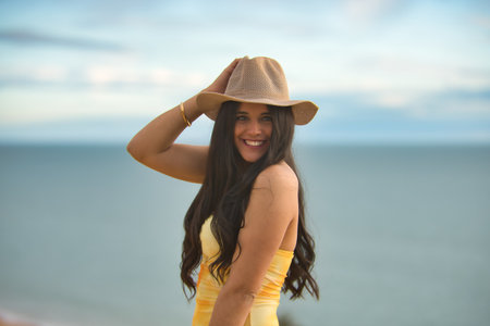 Portrait of young, beautiful, brunette woman, wearing a yellow dress and hat, looking at the camera smiling and happy with the sea in the background. Concept beauty, fashion, beach, sea, sunset.の写真素材