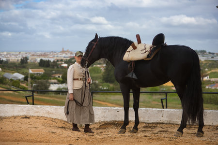 Young woman rider, posing next to her black Spanish thoroughbred horse, which she is holding by the reins. Concept horse riding, horsewoman, horse, dressage.の写真素材