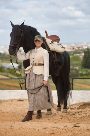 Young woman rider, posing next to her black Spanish thoroughbred horse, which she is holding by the reins. Concept horse riding, horsewoman, horse, dressage. Vertical position.の写真素材
