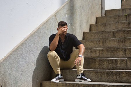 Young, brunette, Hispanic, hip man in black crochet shirt and jeans, depressed and anxious, sitting with his back against a wall on a lonely staircase. Trendy, fashionable, casual concept.の写真素材