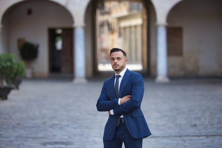 Young, brunette, Hispanic, modern executive, in suit and tie, posing with arms crossed reflecting being a successful man, on a city street.の写真素材