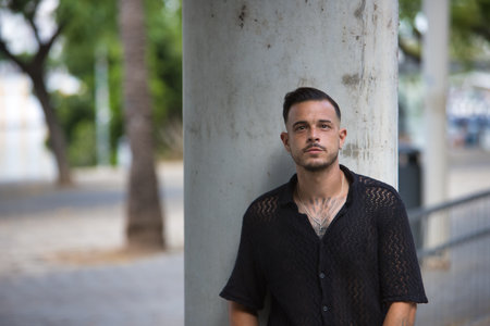 Young, brunette, Hispanic, modern man in black crochet shirt, posing looking at camera with serious gesture and hands in pockets, leaning on a column. Trendy, fashionable, casual concept.の写真素材
