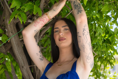 Portrait of a young woman, brunette, tall, slim and very tattooed, wearing a blue top, with a sweet and tender look holding onto a vine of green leaves. Concept of youth, beauty, rebellion, millennialの写真素材