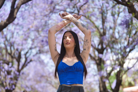 Young woman, brunette, tall, thin and heavily tattooed, wearing a blue top and short leather pants, looking into the distance with her arms crossed above and surrounded by purple bougainvillea trees.の写真素材