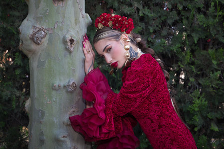 Young woman with red flamenco dress, flowers in her hair and gold jewelry, performing flamenco poses leaning on a tree trunk. Concept dance, art, typical Spanish.の写真素材