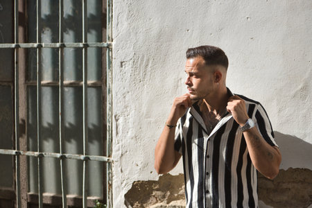 Young, brunette, Hispanic, modern man, wearing crochet shirt with black and white stripes, posing raising shirt collars, receiving sun rays on a white wall.の写真素材