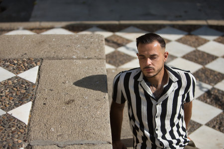 Young, brunette, Hispanic, modern man in crochet shirt with black and white stripes and jeans, eyes closed, relaxed and calm sunbathing. Trendy, fashionable, casual concept.の写真素材
