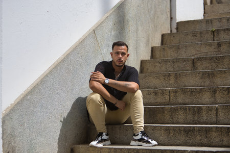 Young, brunette, Hispanic, modern man in black crochet shirt and jeans, smoking sitting with his back against a wall on a lonely staircase. Trendy, fashionable, casual concept.の写真素材