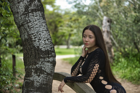 Young woman, dark-haired, beautiful, with pale skin, wearing a black dress, with a distant look, thoughtful and worried, leaning on a wooden railing in an outdoor park.の写真素材