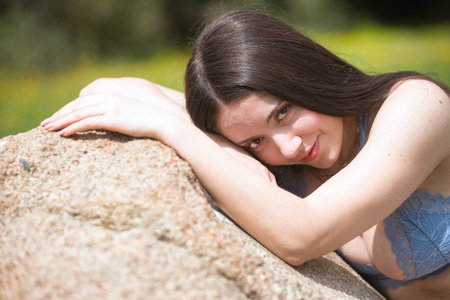 Portrait of a young brunette woman, beautiful, with pale skin, wearing a blue top, shy and reserved, looking at the camera, sitting on a huge stone in an outdoor park.の写真素材