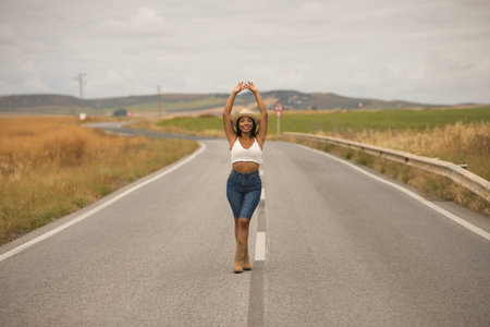 Young, beautiful, Latina brunette woman wearing a white crochet top, jeans, boots, and a hat, with her arms raised, smiling and happy, posing in the middle of a lonely road.の写真素材