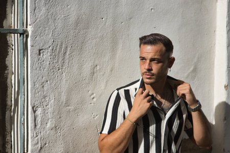 Young, brunette, Hispanic, modern man, wearing crochet shirt with black and white stripes, posing flirtatiously raising shirt collars, receiving sun rays on a white wall.の写真素材