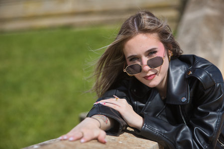 Portrait of young, beautiful, blonde woman, with leather jacket and sunglasses, leaning against a wall looking at the camera over the glasses. Rebellious, indomitable, insurgent concept.の写真素材