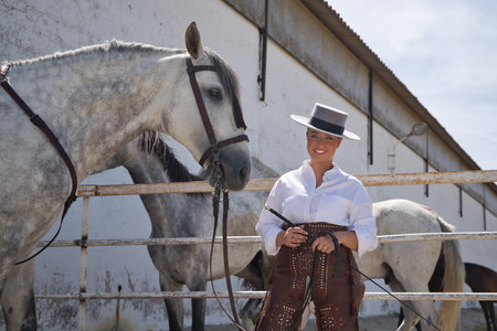 Young, beautiful, blonde woman in white shirt, leather chaps, hat and a riding crop in her hands, posing looking at camera in a stable surrounded by horses. Concept animals, riding, horses, sport.の写真素材