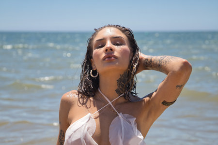 Portrait of a young, beautiful, Latina woman with tattoos, wearing a white dress, wet, with one hand on her head, posing stylishly and glamorously in the sea on a sunny beach day. Concept: vacation.の写真素材