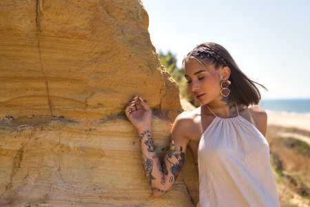 Young, Latina woman, heavily tattooed, wearing a white dress, eyes closed, relaxed and alone, leaning against a cliff by the beach surrounded by nature. Concept: femininity, beauty, fashionの写真素材