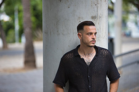 Young, brunette, Hispanic, modern man in black crochet shirt, posing with serious and worried gesture and hands in pockets, leaning on a column. Trendy, fashionable, casual concept.の写真素材