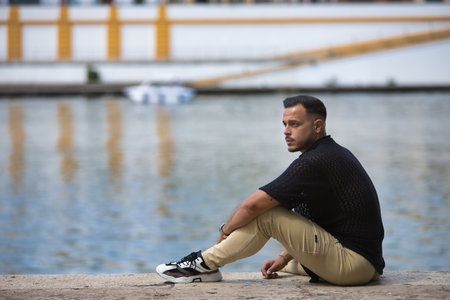 Young, brunette, Hispanic, modern man in black shirt and jeans, relaxed and calm, looking at infinity, sitting on the ground by the river. Trendy, fashionable, casual concept.の写真素材