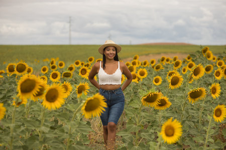 Young, attractive, Latina brunette woman wearing a white crochet top, jeans, and a hat, smiling and happy as she walks through a field of sunflowers. Concept: nature, agriculture, countryside.の写真素材
