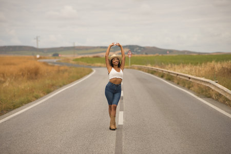 Young, beautiful, Latina brunette woman wearing a white crochet top, jeans, boots, and a hat, with her arms raised, smiling and happy, posing in the middle of a lonely road.の写真素材