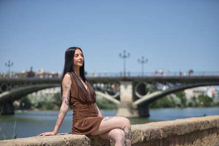 Young woman, brunette, tall, slim and very tattooed, with brown dress, looking to infinity sitting by the river with the Triana bridge in the background. Concept of youth, millennial.の写真素材