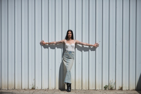Young woman, brunette, tall, slim and very tattooed, with white top, denim skirt and boots, posing looking at the camera, with open arms leaning on a white metal wall. Concept youth, beauty.の写真素材