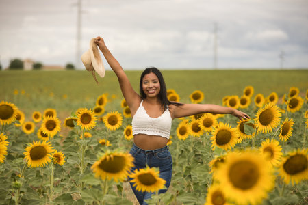 Young, attractive, Latina brunette woman wearing a white crochet top, jeans, and a hat in her hand, smiling happily with her arms raised in a field of sunflowers. Concept: nature, agriculture.の写真素材