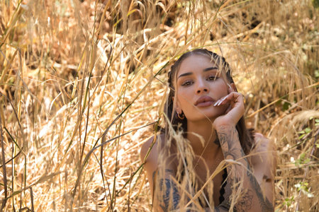 Portrait of a young, beautiful, Latina woman with tattoos, wearing a white crochet dress, intimate and tender, connected to nature and ecology, sitting on the ground surrounded by dry grasses.の写真素材