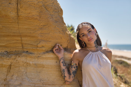 Young, beautiful, Latina woman, heavily tattooed, wearing a white dress, with a dreamy look, leaning on a cliff next to the beach surrounded by nature. Concept: femininity, beauty, fashion, nature.の写真素材
