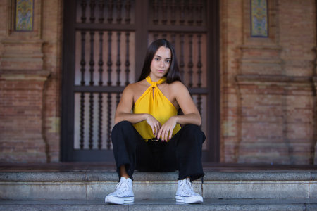 Young woman, brunette, wearing a yellow shirt, black jeans, and white sneakers, with a strong, penetrating gaze, sitting on some steps. Concept: youth, trendy, fashion, modern.の写真素材