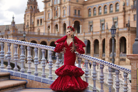 Young, beautiful, blonde woman in red flamenco dress, flowers in her hair and gold jewelry, performing flamenco poses on a nice bridge in the square of Spain in Seville. Dance concept, art, typical.の写真素材