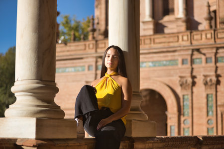 Young woman, brunette, Latina, wearing a yellow shirt, sitting, leaning against a marble column, resting, basking in the sun in Spain square in Seville. Concept: tourism, travel, charming places.の写真素材