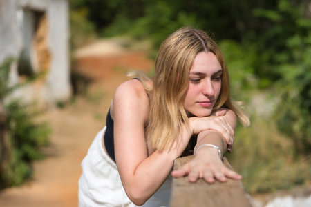 Portrait of young, beautiful, blonde woman in black T-shirt and white pants, looking distracted, leaning on a wooden railing in an outdoor park. Concept beauty, youth, millennial.の写真素材
