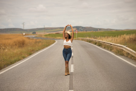 Young, beautiful, Latina brunette woman wearing a white crochet top, jeans, boots, and a hat, with her arms raised, smiling and happy, posing in the middle of a lonely road.の写真素材