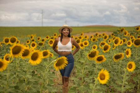 Young, attractive, Latina brunette woman wearing a white crochet top, jeans, and a hat, smiling and happy as she walks through a field of sunflowers. Concept: nature, agriculture, countryside.の写真素材