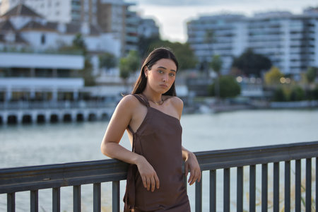 Young woman, brunette, blue eyes, Hispanic, wearing an elegant brown chiffon dress, posing with a fixed, penetrating gaze, leaning on a metal railing. Concept: model, beauty, fashion.の写真素材