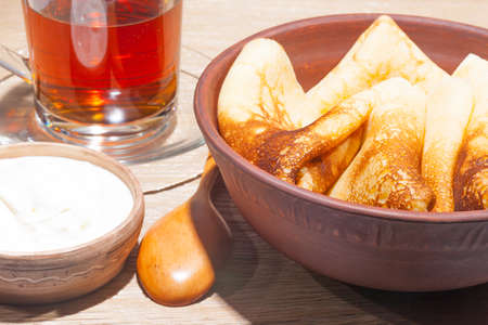 traditional oiled food in vintage style: pancakes with sour cream in and a clay plate and tea in a glass cup with a wooden spoon on an oak table
の写真素材