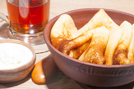 traditional oiled food in vintage style: pancakes with sour cream in and a clay plate and tea in a glass cup with a wooden spoon on an oak table
の写真素材