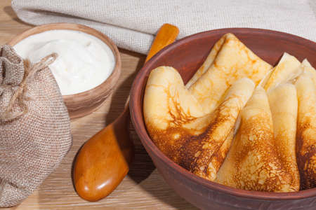 traditional oily food in vintage style: pancakes with sour cream in and a clay dish with a wooden spoon on an oak tableの写真素材