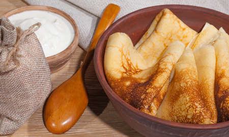 traditional oily food in vintage style: pancakes with sour cream in and a clay dish with a wooden spoon on an oak tableの写真素材