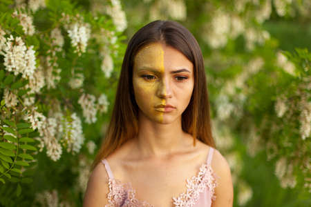 close-up portrait of a beautiful young girl in around white acacia in the spring half face golden make-upの写真素材