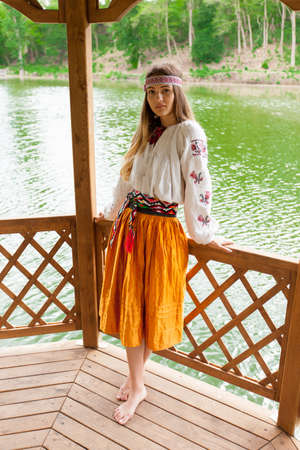 Beautiful Ukrainian woman dressed in embroidery in a wooden summer altanka on a lakeの写真素材