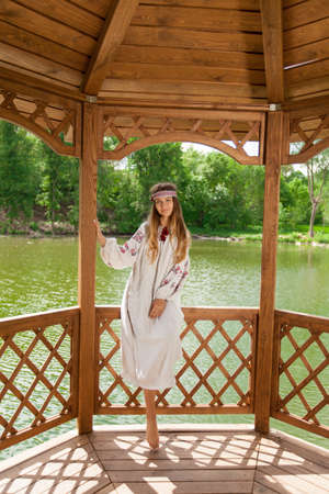 Beautiful Ukrainian woman dressed in embroidery in a wooden summer altanka on a lakeの写真素材