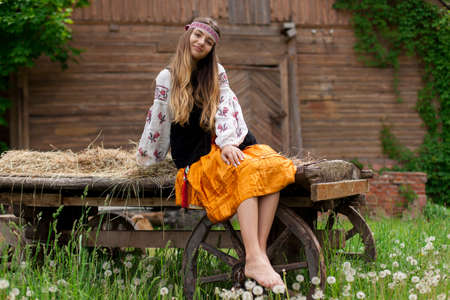 Beautiful slender Ukrainian woman in embroidery on the background of a wooden frame of a barn in the village sits on a wooden cartの写真素材