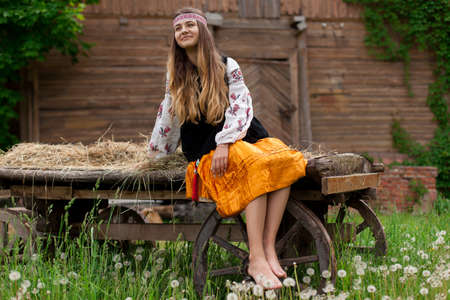 Beautiful slender Ukrainian woman in embroidery on the background of a wooden frame of a barn in the village sits on a wooden cartの写真素材
