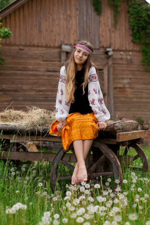 Beautiful slender Ukrainian woman in embroidery on the background of a wooden frame of a barn in the village sits on a wooden cartの写真素材